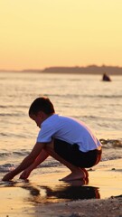 Wall Mural - Boy plays in sea water in the evening. Side view of a boy in white t-shirt and shorts playing on sea shore at sunset. Vertical video