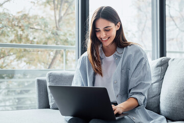 Concentrated millennial girl sit on couch working on laptop browsing internet at home during lazy weekend, focused young woman freelancer busy using computer surfing wireless web shopping online.
