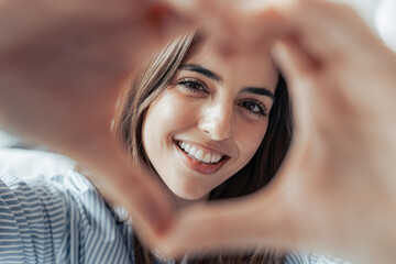 Beautiful woman showing heart sign with folded fingers smiling looking at camera, head shot portrait. Attractive optimistic young female demonstrate symbol of love feels happy posing alone at home.