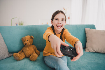 Wall Mural - Portrait of little schoolgirl sit couch hold controller enjoy play video games flat indoors