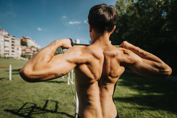 Wall Mural - Fit young man performing a resistance band workout outdoors, showcasing a strong, muscular back in a sunny park setting.