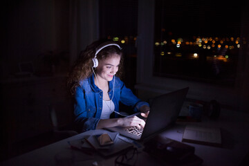 Wall Mural - Beautiful university student is writing her thesis on a laptop at night, sitting at home at table, focusing on her research. Preparing for final exam, studying.