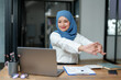 © crizzystudio - Modern office Muslim businesswoman wearing hijab sits and works with laptop online for a long time from fatigue and arm pain, meditate and stretch to relax. Office syndrome concept.