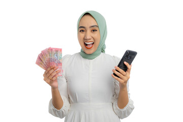 Wall Mural - Excited young Asian woman in green hijab and white blouse holding mobile phone and money with excitement isolated on white background