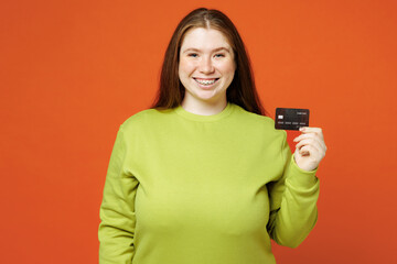 Wall Mural - Young fat ginger chubby plus size woman wear green sweatshirt casual clothes hold in hand mock up of credit bank card isolated on red orange background studio portrait Lifestyle body positive concept