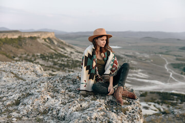 Wall Mural - serene woman in the mountains sitting on a rock with a hat and blanket on her shoulders