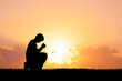 © STOCK PHOTO 4 U - Silhouette of a Person Kneeling in Prayer at Sunset with a Vibrant Orange Sky and Clouds in the Background, Capturing a Moment of Solitude and Reflection in Nature