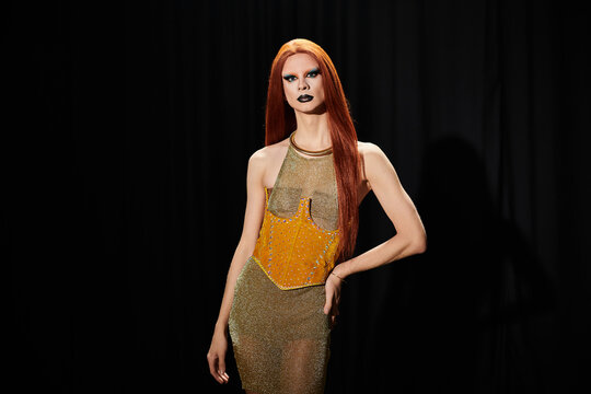 A drag queen in a gold sequined dress poses against a black backdrop.