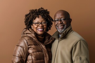 Wall Mural - Portrait of a glad afro-american couple in their 40s donning a durable down jacket over soft brown background