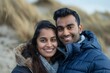 © Markus Schröder - Portrait of a grinning indian couple in their 30s donning a durable down jacket while standing against sandy beach background