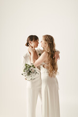 Wall Mural - Two brides, dressed in white, stand close and embrace, looking into each others eyes. The bride on the left holds a bouquet of white flowers.