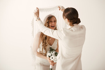 Wall Mural - Two brides in white wedding attire share a joyful moment during their wedding ceremony, as one playfully unveils the others veil.