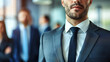© Domingo - A confident businessman in a suit and tie stands in focus, with blurred colleagues in the background, suggesting leadership, teamwork, and professional environment in a modern office setting.