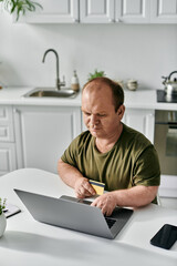 Wall Mural - A man with inclusivity sits at a kitchen table, using his laptop and a credit card for online shopping.