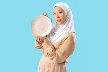 Poster - Young Asian woman in hijab with plate on blue background