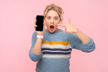 Portrait of shocked surprised blonde woman wearing sweatshirt pointing at smartphone with empty display, copy space for promotion. Indoor studio shot isolated on light pink background.