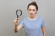 © khosrork - Portrait of woman wearing striped T-shirt looking through magnifying glass, finding out something, magnifies small details to see, inspecting. Indoor studio shot isolated on gray background.