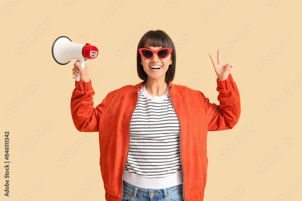 Young woman with megaphone showing victory gesture on beige background