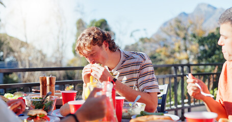 Canvas Print - Friends, man and eating hamburger outdoor on terrace with hunger, reunion and social event in summer. People, barbecue and lunch on patio with food, feast or celebration with bonding and drinks