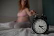 © Михаил Решетников - Caucasian pregnant woman sits on the bed and suffers from insomnia. Alarm clock in the foreground.