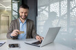 © Liubomir - Smiling businessman using laptop and credit card for online payment in modern office. Concept of e-commerce, online transactions, and business technology.