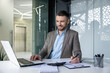 © Liubomir - Confident businessman smiling while working on his laptop and taking notes in a sleek, modern office environment.