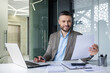 © Liubomir - Smiling businessman holding a document while working in a modern office. Professional environment with a laptop, papers, and a pen.