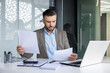 © Liubomir - Businessman examining papers, multi-tasking at an office desk with a laptop, looking concerned or focused.