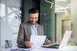 © Liubomir - Smiling businessman reviewing documents and working on a laptop in a modern office setting. Professional, productive environment with positive and successful outcomes.