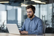 © Tetiana - A smiling young man in a blue shirt is sitting at a desk in a modern office, wearing a headset, working and studying at a laptop