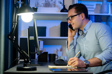 Wall Mural - Working businessman calling phone with customer, typing brief message at neon workplace, planning online business using information project at night time surrounded by stationary on desk. Sellable.