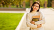 © Prostock-studio - Asian young woman, wearing glasses and earbuds, walks through a sunny university campus. She holds several books and appears content.