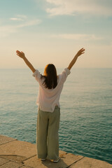 Young woman hands up seen from behind standing on the pier while looking at the sea, relax, joy and freedom, vacation and summer lifestyle, powerful nature