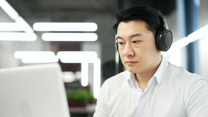 Serious asian IT specialist coder in wireless headphones typing on laptop computer while sitting at workplace in business office. Focused confident male developer in shirt works on a project. Close up