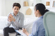 © Prostock-studio - Unhappy man sitting on a couch engaged in conversation with a therapist in a office setting. The man appears focused and attentive while the therapist listens and provides guidance.