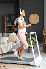 Poster - Sporty young African-American woman with headphones running on treadmill in living room