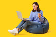 © Pixel-Shot - Pretty young woman with laptop sitting on bean bag chair against yellow background