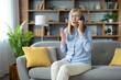 © Liubomir - Elderly woman sitting on a couch, expressing frustration while talking on the phone with customer support in living room.