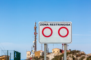 Telecoms masts and buildings on top of the mountain Puig de la Llorenca on a clear sunny day overlooking Cumbre Del Sol, Spain with a Spanish warning sign, English translation  RESTRICTED AREA