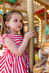 Adorable little brunette girl in summer red-white dress at amusement park having a ride on the merry-go-round. Child girl has fun outdoor on sunny summer day. Entertainment concept