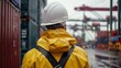 © WITTAYA  ANGMUJCHA - worker in a hard hat and yellow jacket in the rain in a cargo port, view from the back