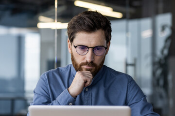 Close-up photo of a serious young businessman, programmer and developer sitting in the office at the desk, thoughtfully and intently looking at the laptop screen