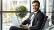 © VK Studio - A young businessman in a sharp suit, smiling while working on a laptop in a bright office space with large windows and green plants.
