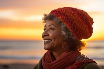Wall Mural - Portrait of a glad afro-american woman in her 60s donning a warm wool beanie while standing against vibrant beach sunset background