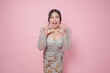 © Reezky - Cheerful Asian woman wearing modern kebaya dress feeling surprised and shouting wow, isolated by pink background.