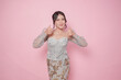 © Reezky - Excited Indonesian woman in modern kebaya dress gives thumbs up hand gesture of approval, isolated by pink background.