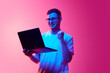 © master1305 - Young man in glasses and white t-shirt, standing with laptop and gesturing, expressing success against pink background in neon light. Concept of human emotions, facial expression, lifestyle