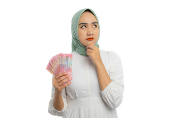 Wall Mural - Pensive young Asian woman in green hijab and white blouse touching his chin, holding money, looking up with doubtful expression isolated on white background