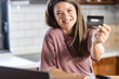 © lordn - A young cheerful woman is buying groceries by ordering them online and paying for them with a card