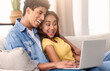 © Prostock-studio - A teen couple is sitting on a couch together, using a laptop and laughing. The guy has his arm around the girl and they both appear to be enjoying each others company.